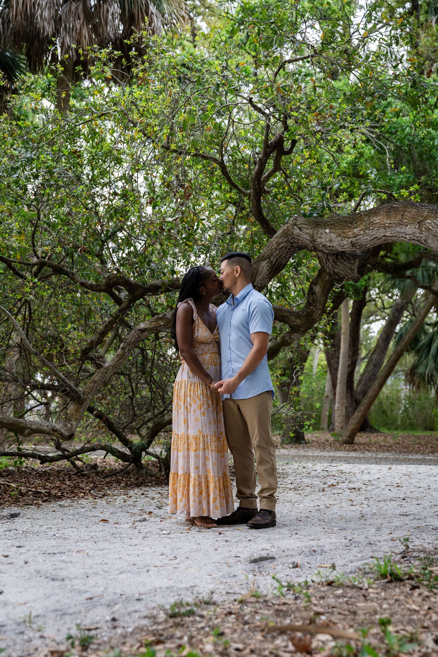 A couple shares a romantic kiss while holding hands on a sandy path surrounded by lush green trees.