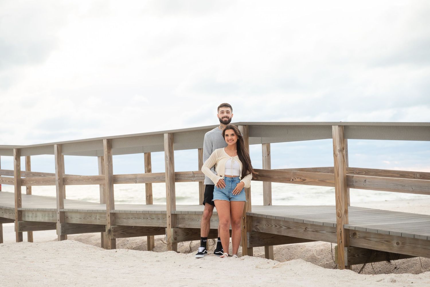 Couple posing together on a wooden beach boardwalk with sandy shores and cloudy skies in the background.