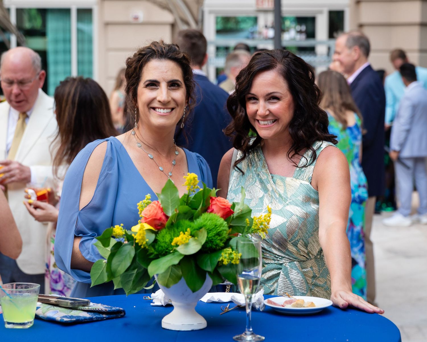 Two women smile at outdoor event beside bright floral centerpiece on blue-draped table.