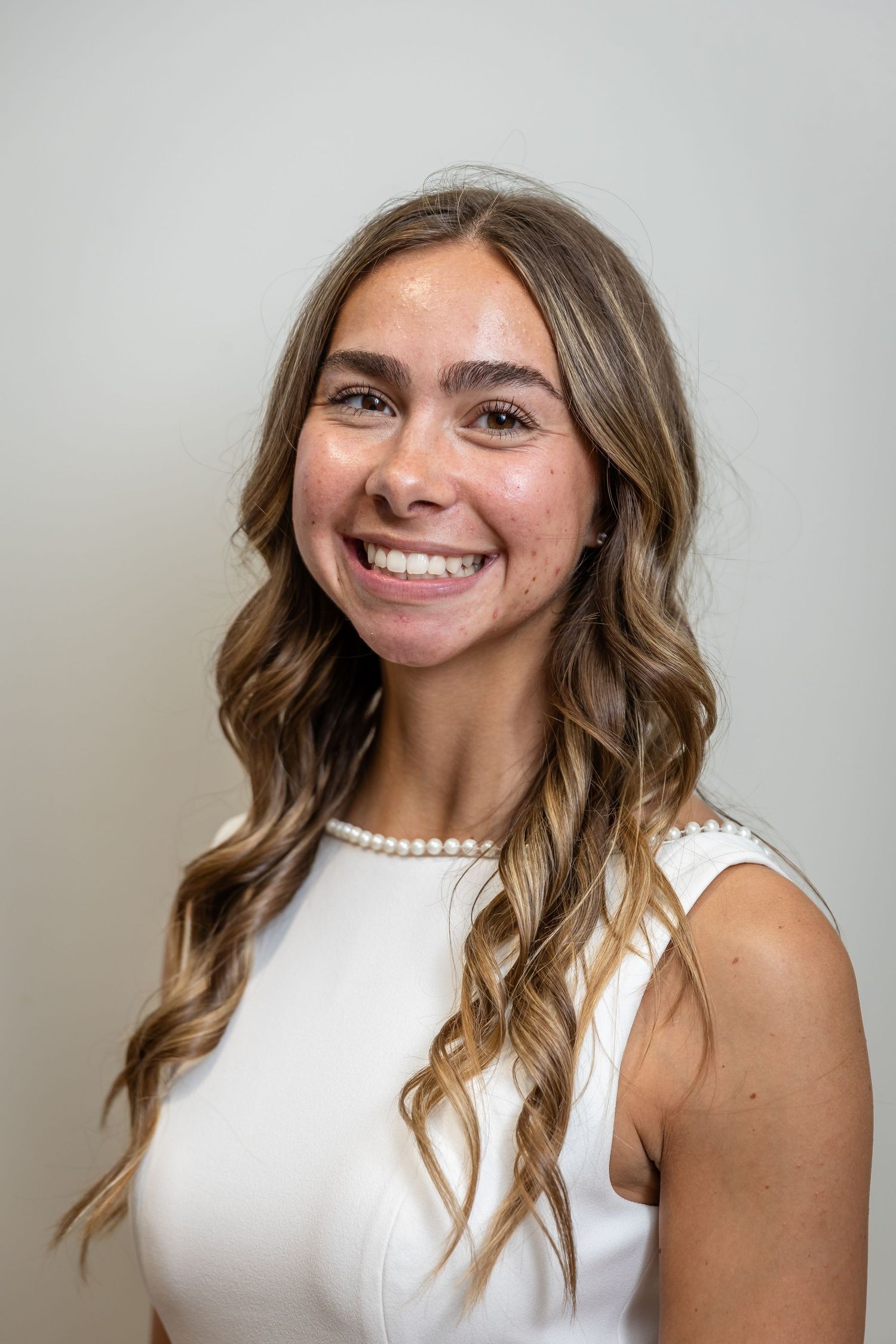 Young woman with long wavy hair smiling, wearing a white sleeveless top and pearl necklace, professional headshot.