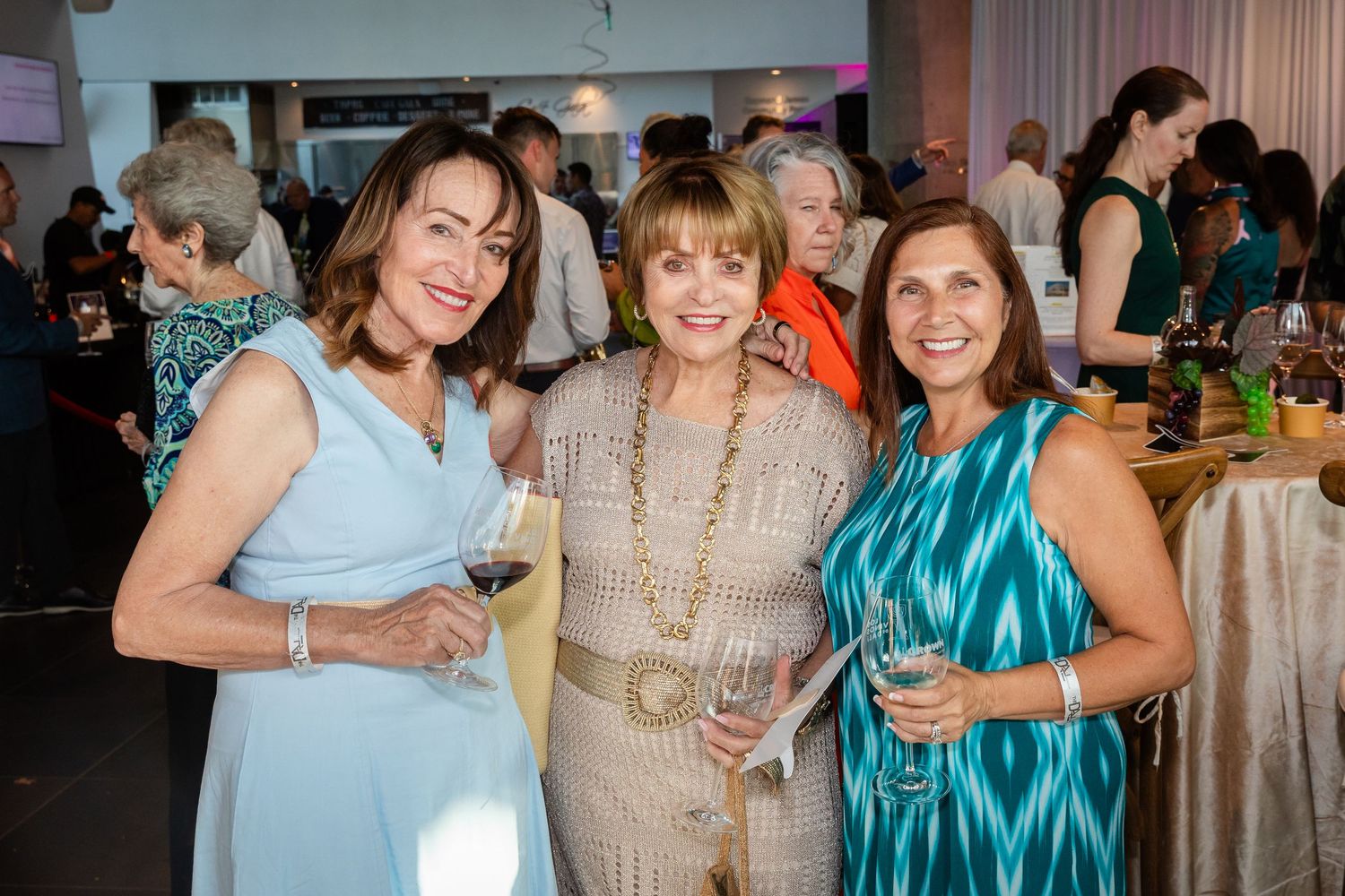 Three elegantly dressed women smiling and holding wine glasses at a formal indoor social event.