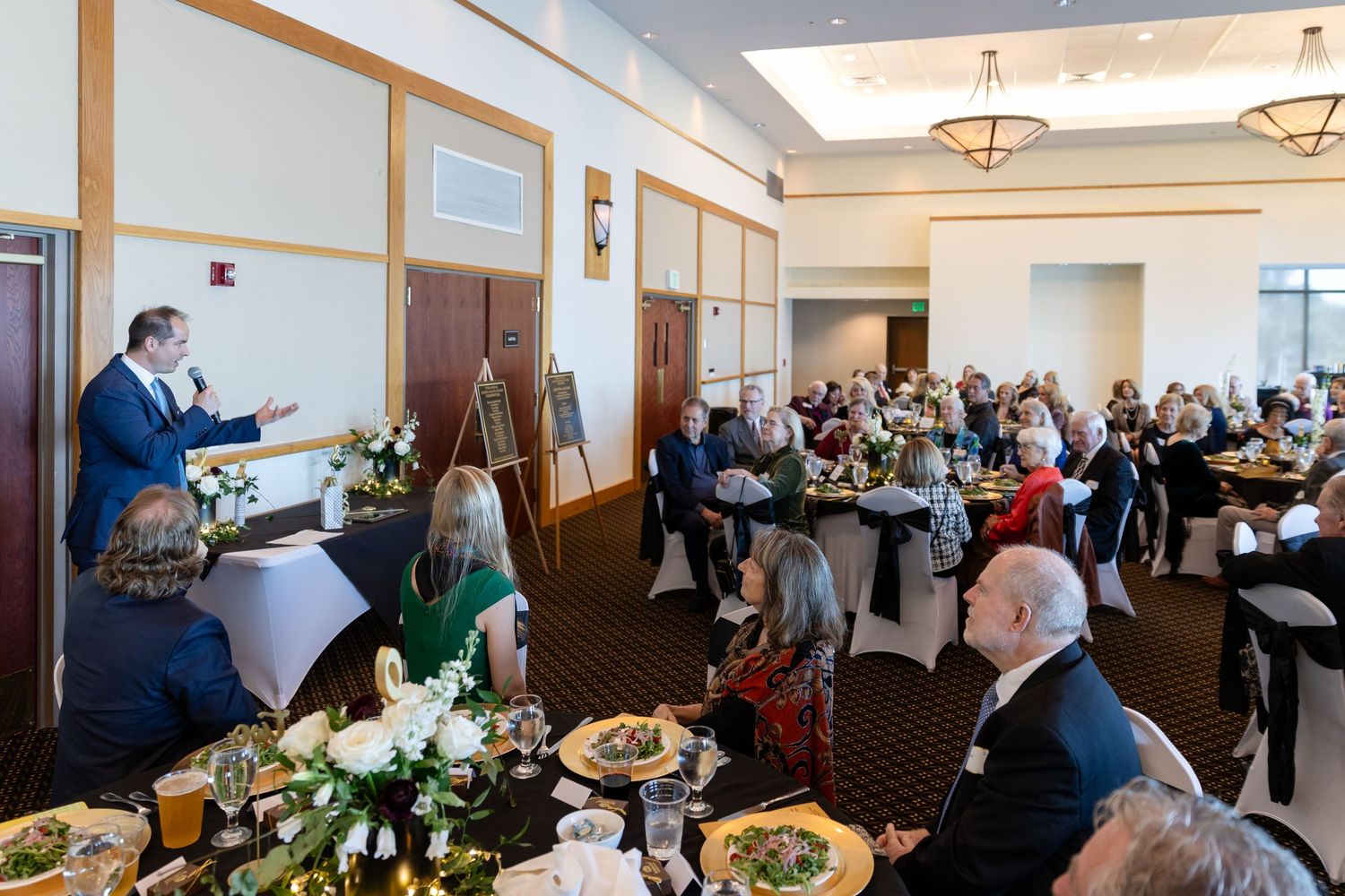 Speaker addresses guests seated at decorated tables during an elegant formal luncheon event in a banquet hall.