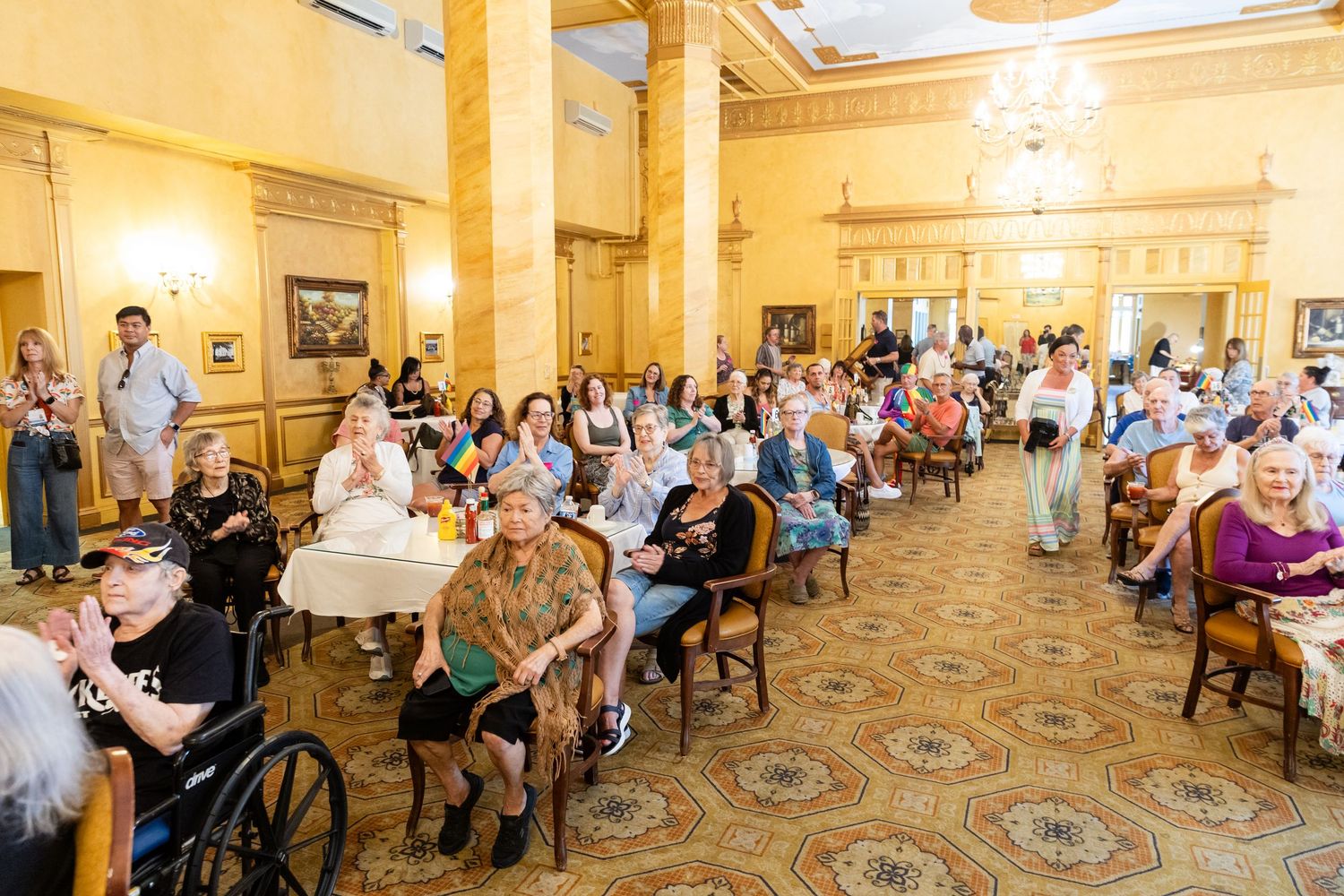 Elderly residents and guests gathered in an ornate golden ballroom for a festive community celebration event.