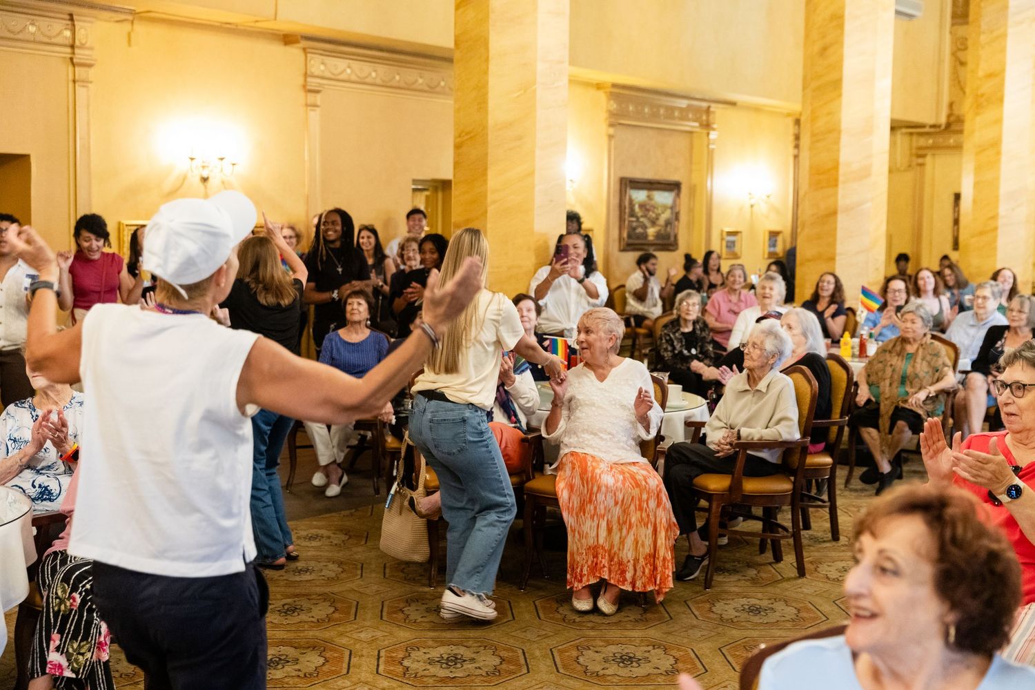 Diverse crowd of seniors clapping and dancing at a lively indoor Pride celebration event in an elegant hall.