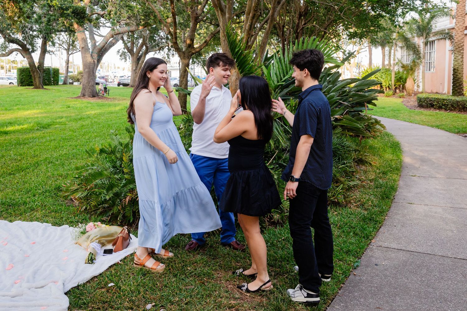 Four teenagers socializing outdoors on a sunny day, dressed in formal attire on a green lawn near tropical plants.