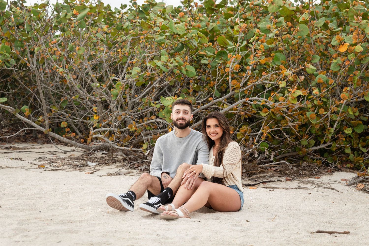 A young couple sits together on sandy beach, smiling, with lush green shrubs in the background.