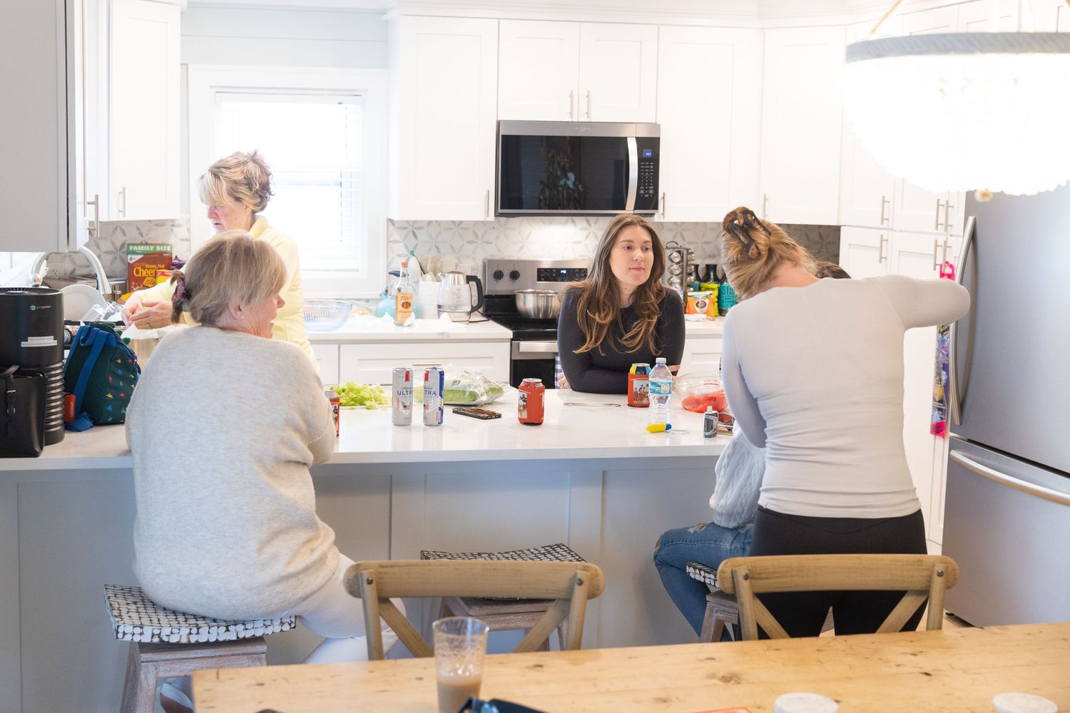Four women gather around a white kitchen island, socializing in a bright modern kitchen with stainless steel appliances.