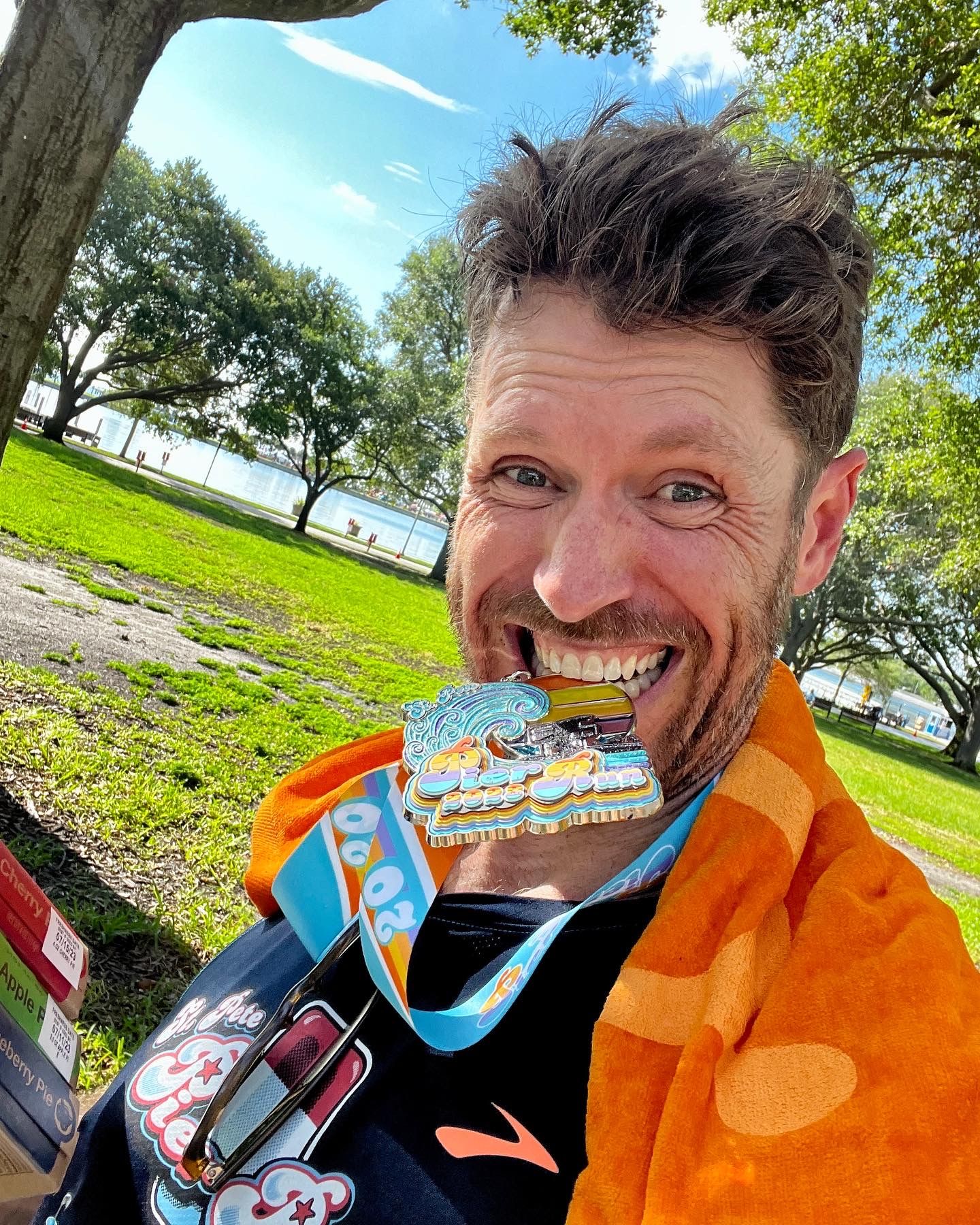 Man smiling with race finisher medals around his neck wearing an orange towel outdoors near a park.