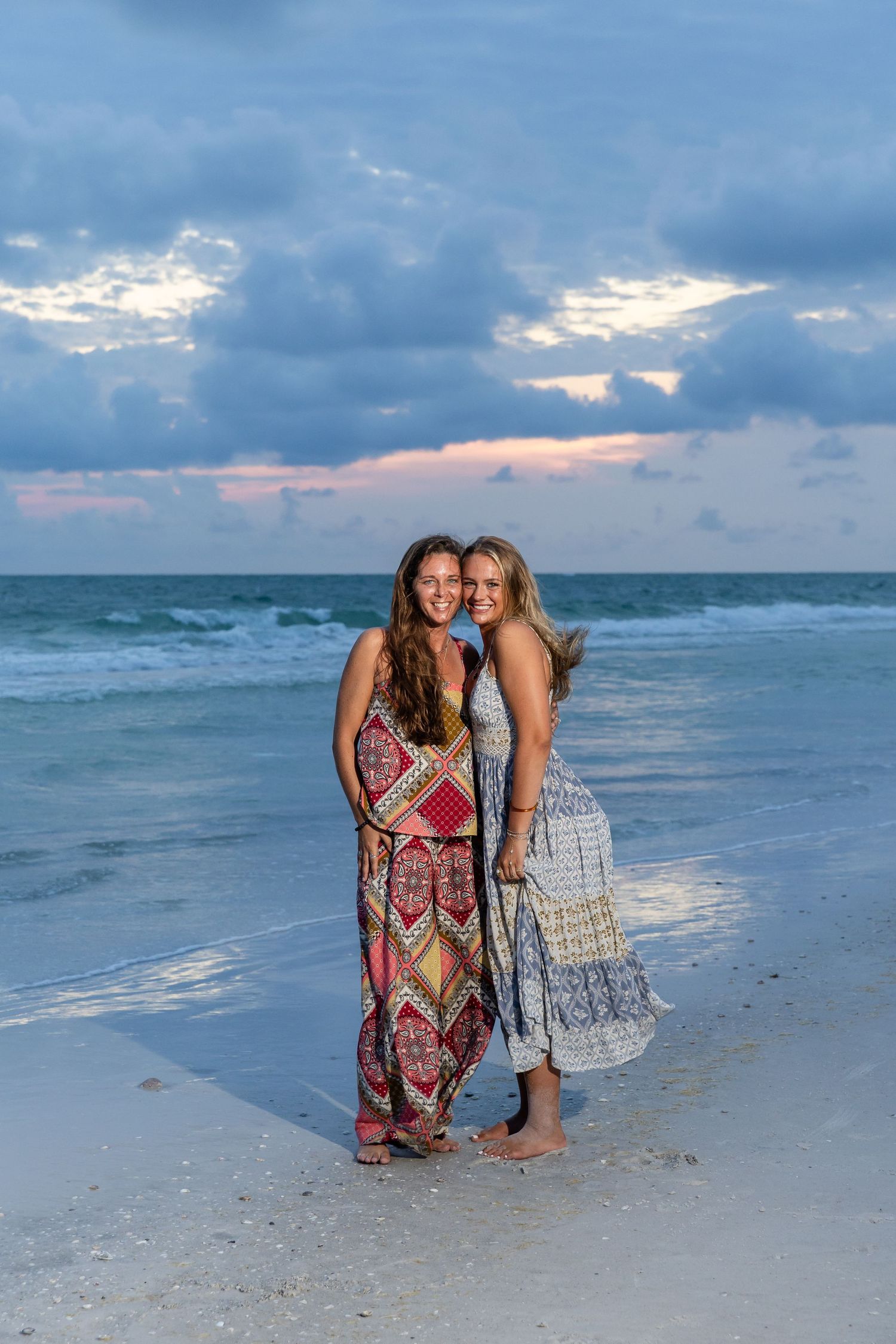 Two smiling women in summer dresses posing together on beach at dusk with ocean waves behind.
