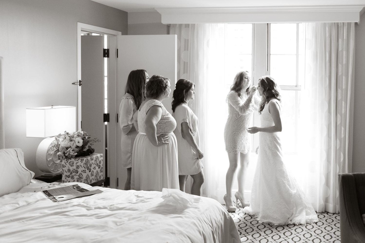 Black and white photo of bridesmaids in robes helping the bride into her wedding gown by a window.