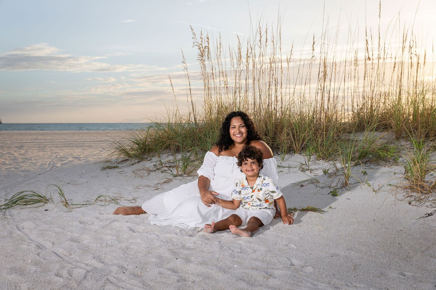 A mother and child sit together on white sand dunes at sunset, both wearing white outfits with sea oats behind them.