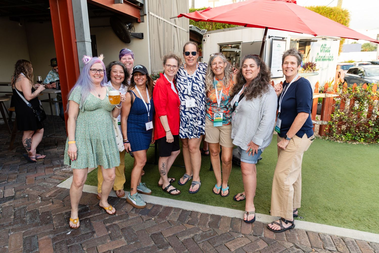 Group of women smiling together outdoors at a brewery event on a sunny day.