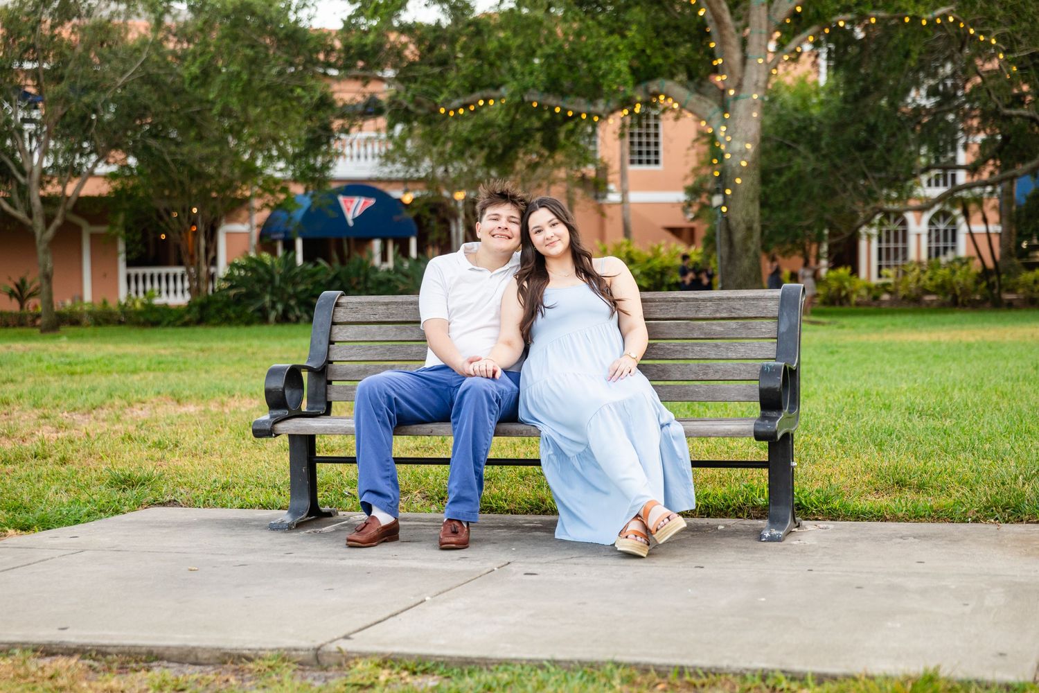 Couple sitting together on a park bench outdoors, woman in light blue dress, man in jeans, string lights in background.