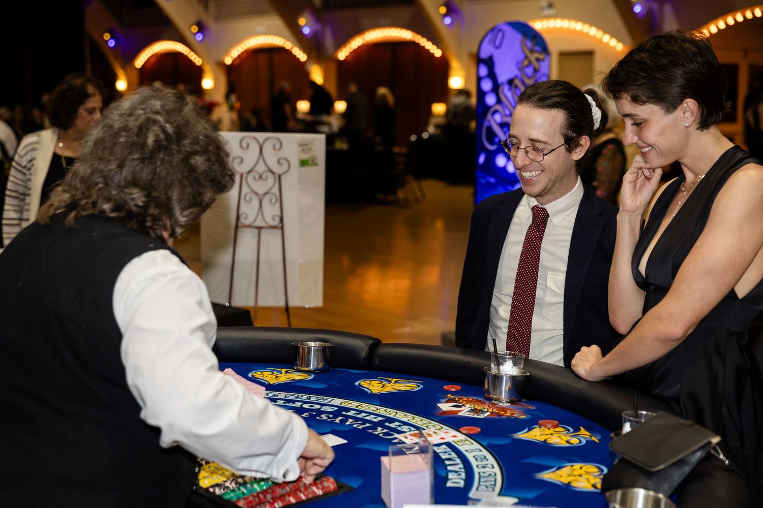 Casino blackjack table with dealer and two elegantly dressed guests playing cards at an indoor event.
