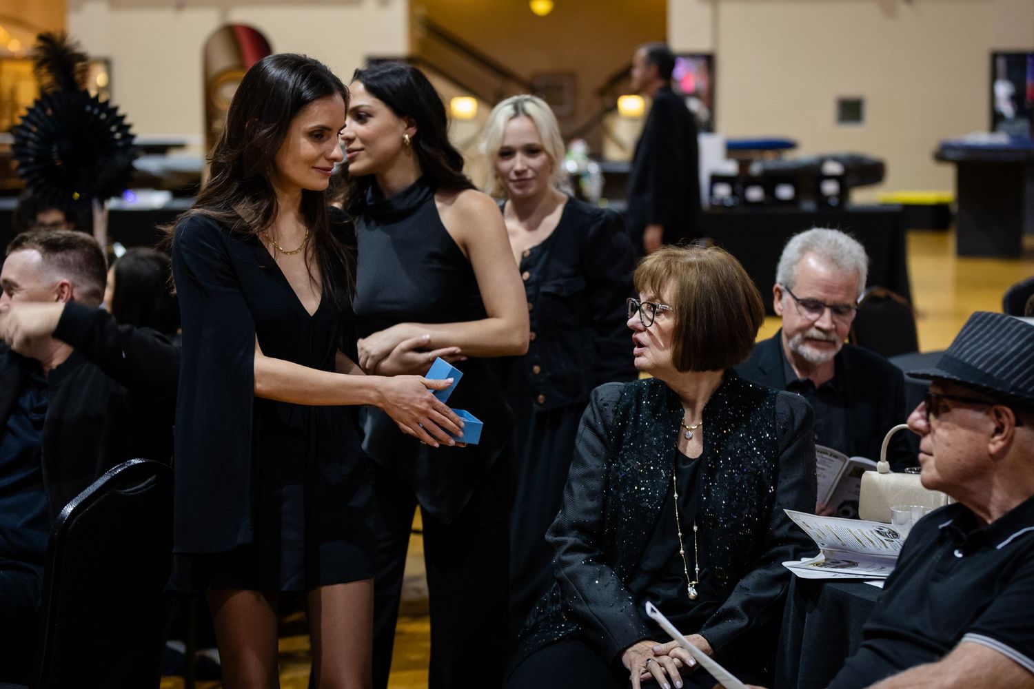 Elegantly dressed guests in black attire socialize and interact at an upscale evening gala event.