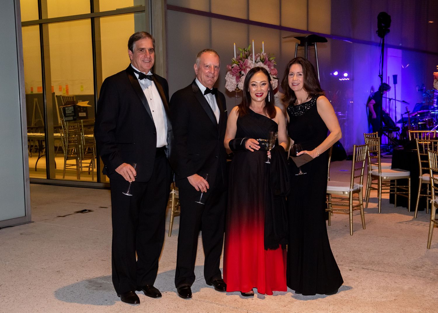 Two couples in formal black tie attire pose together smiling at an elegant evening gala event.