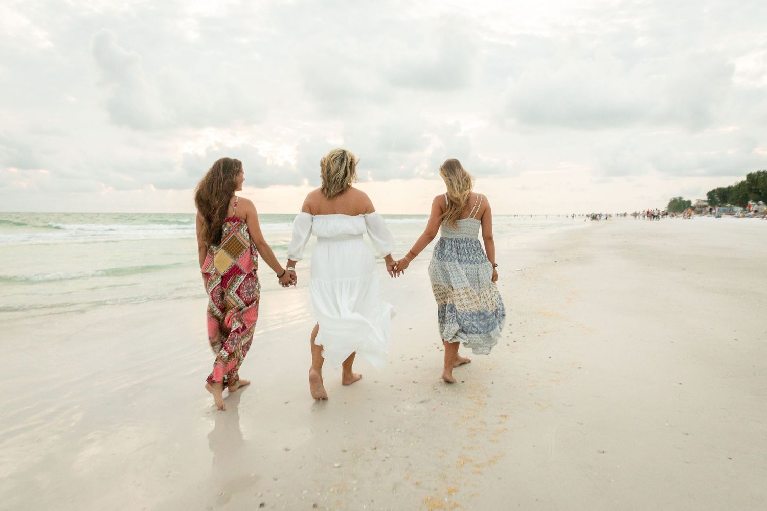 Three women in colorful dresses hold hands walking along a sandy beach with ocean waves.