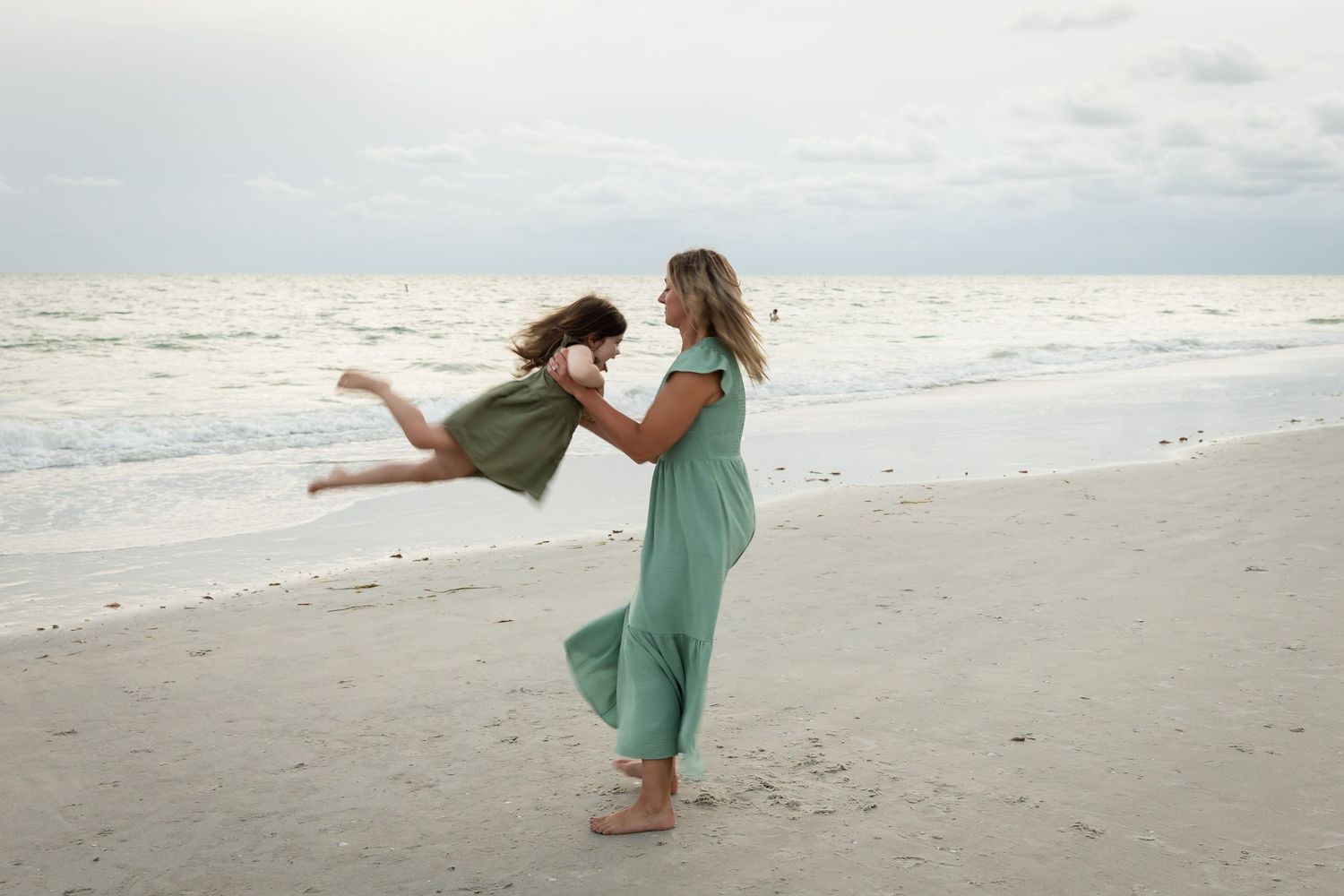 Mother in green jumpsuit twirling young daughter on sandy beach, both wearing matching outfits by the ocean.