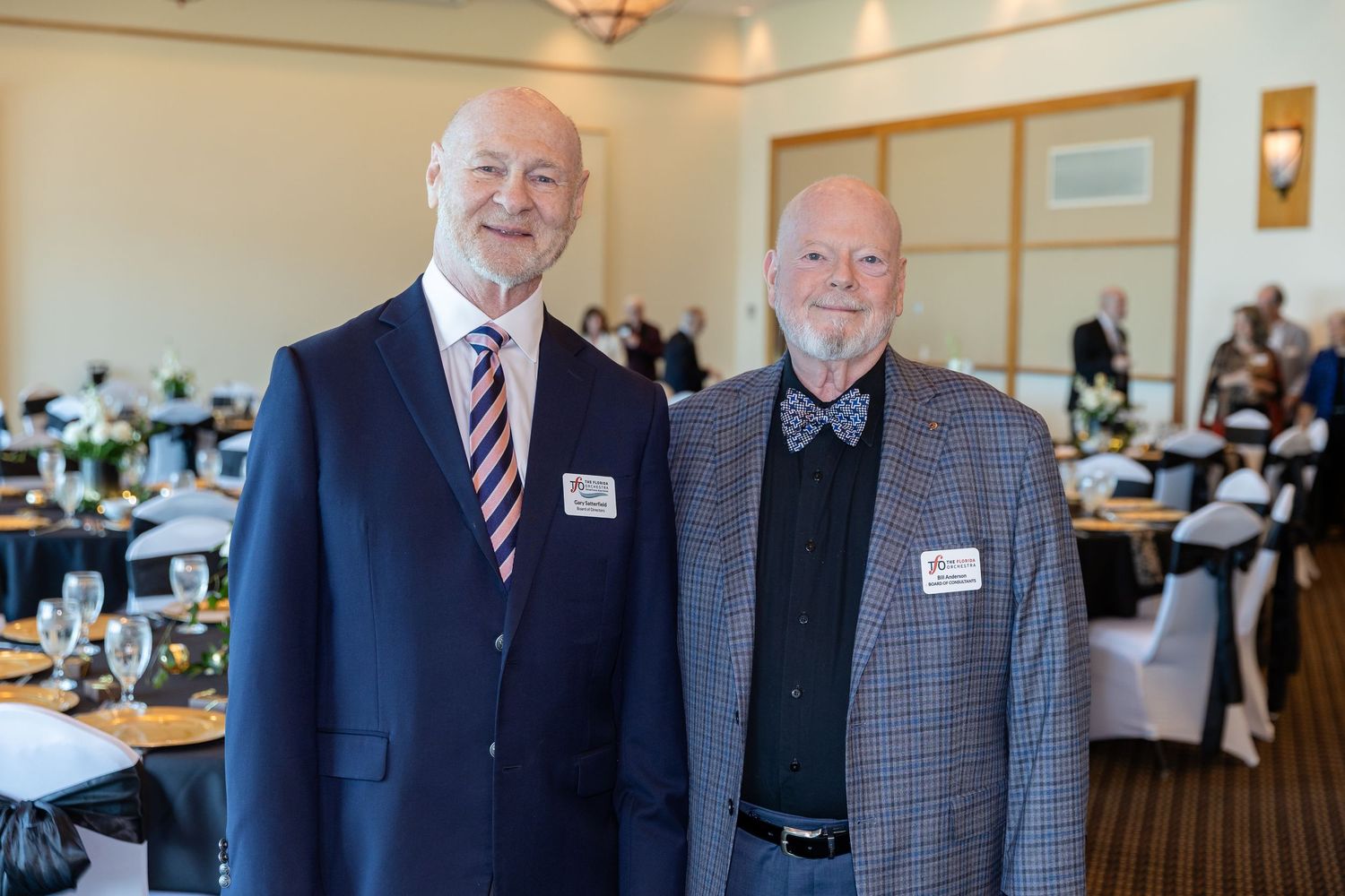 Two senior men in suits smiling at a formal banquet event with elegantly set tables in the background.