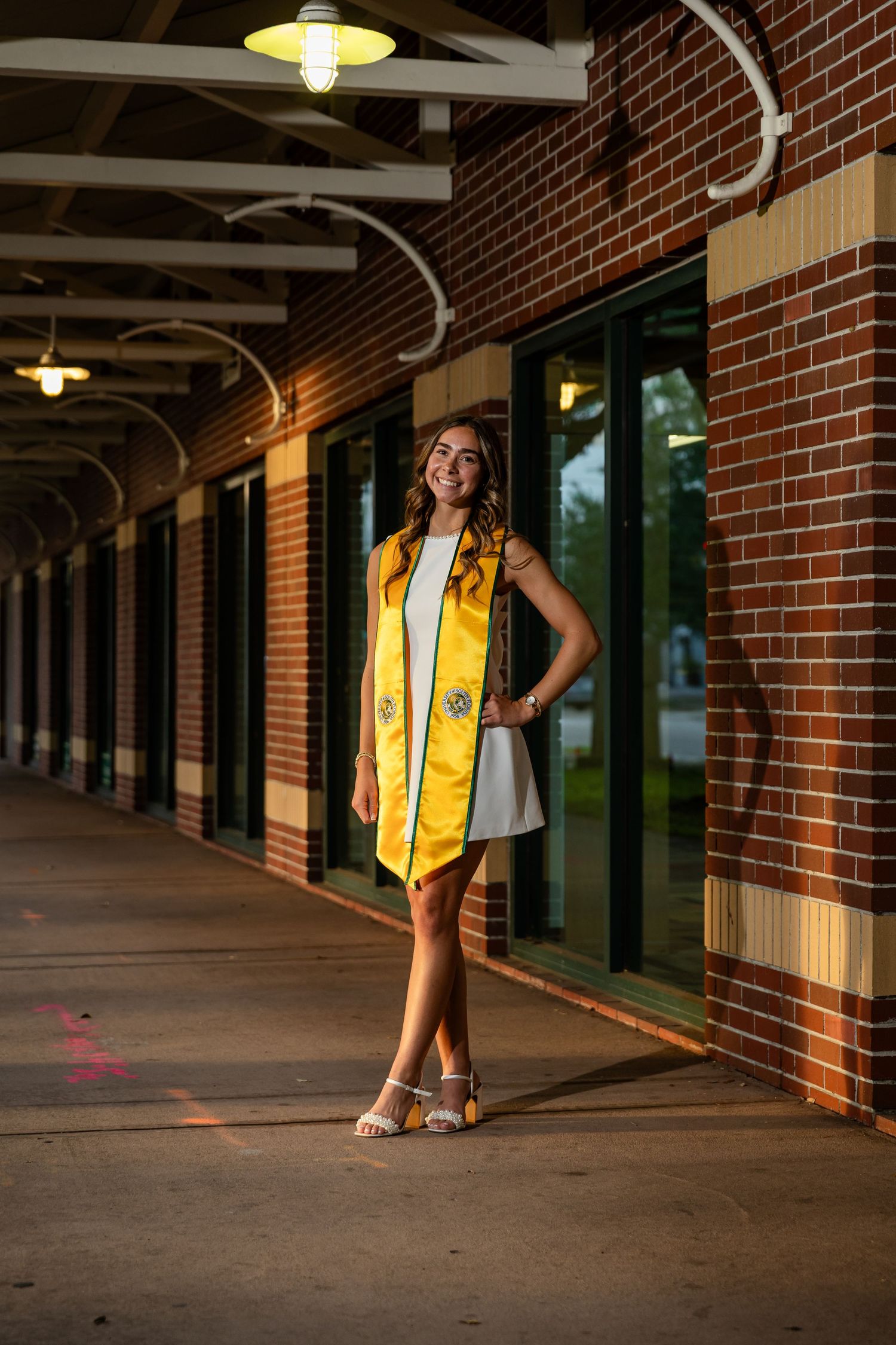 Smiling graduate wearing a yellow stole poses in a white dress along a brick building corridor.