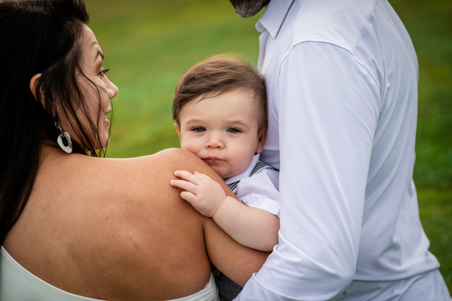 A close-up of a baby resting on parents shoulders, gazing at the camera with bright blue eyes outdoors.