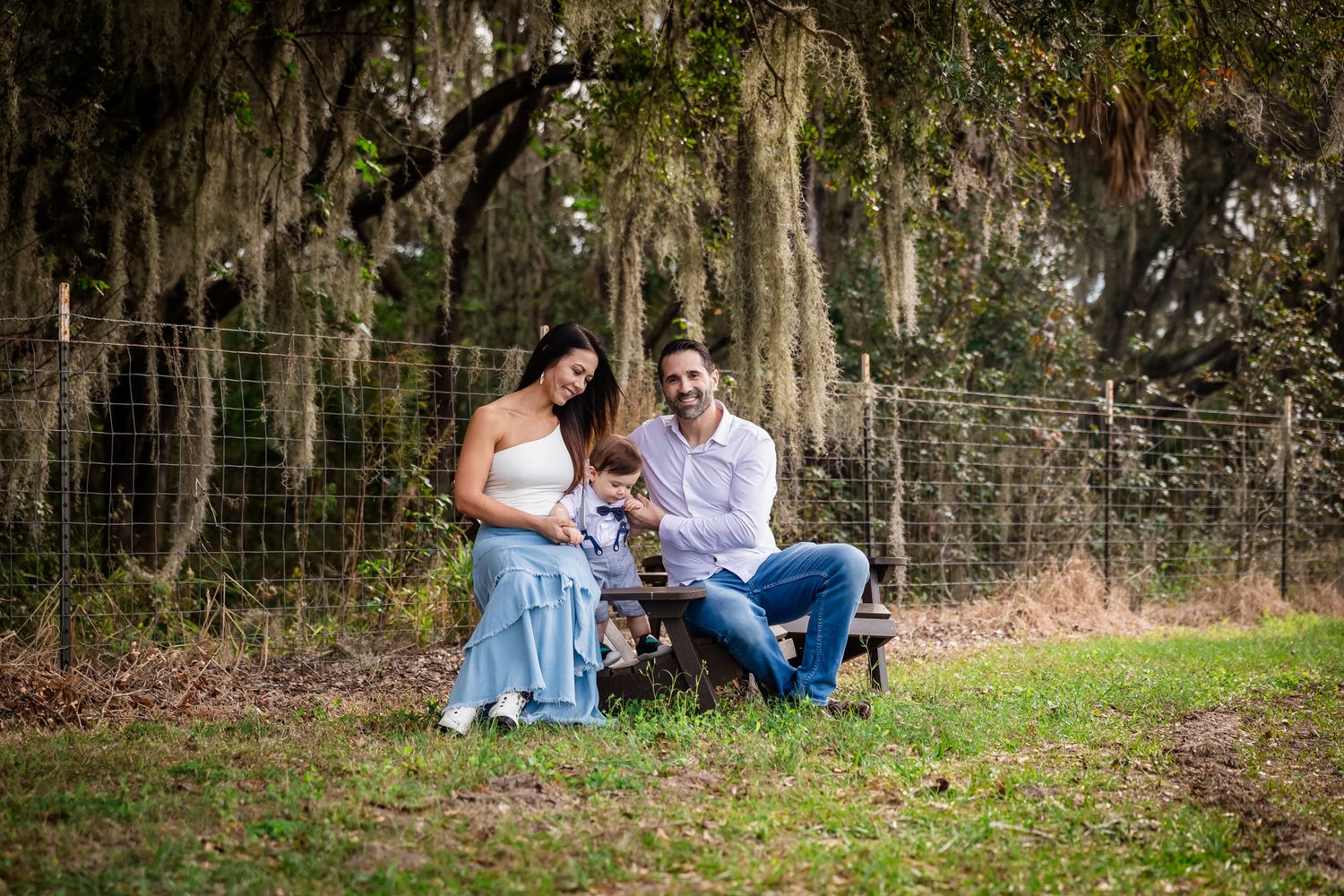 Happy family sitting outdoors under Spanish moss-draped oak trees, parents smiling with baby on rustic bench.