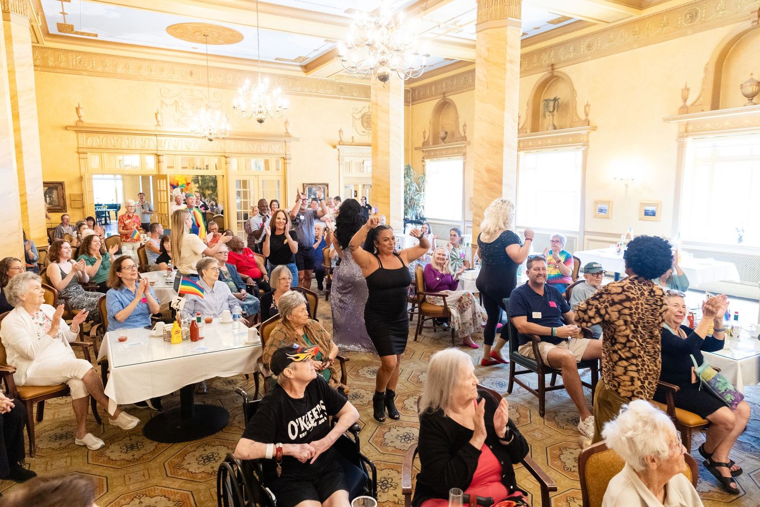 Large crowd of diverse guests celebrating Pride in a grand ballroom with performers and festive decorations throughout the room.