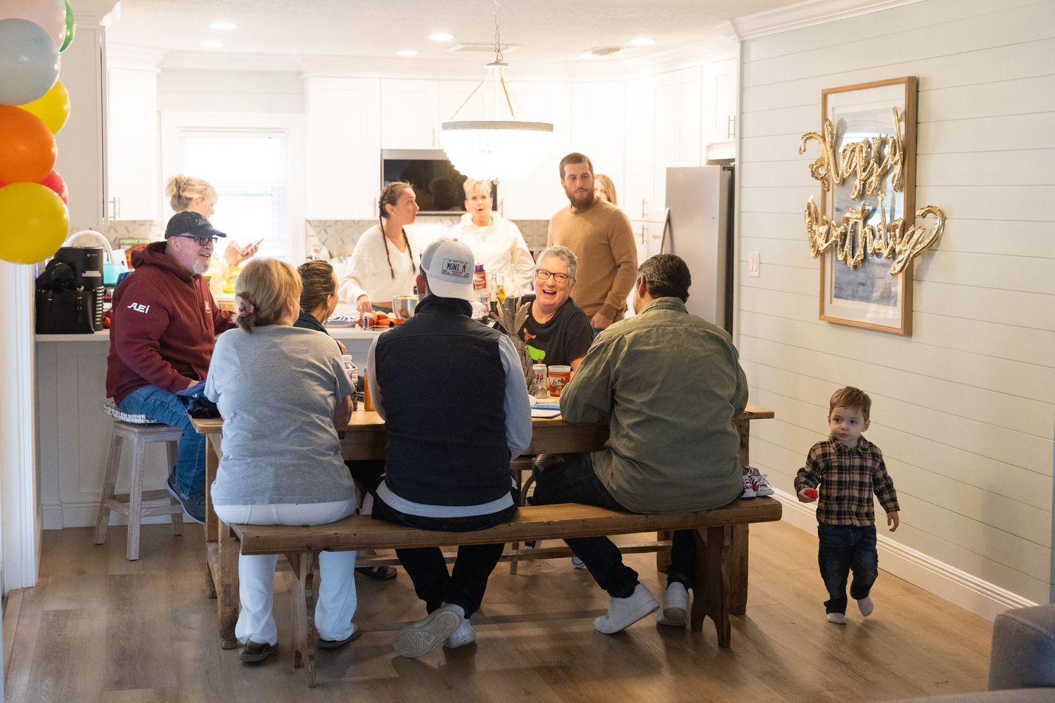 Family gathering around a rustic dining table in a modern kitchen, with a child standing nearby and colorful balloons in the background.