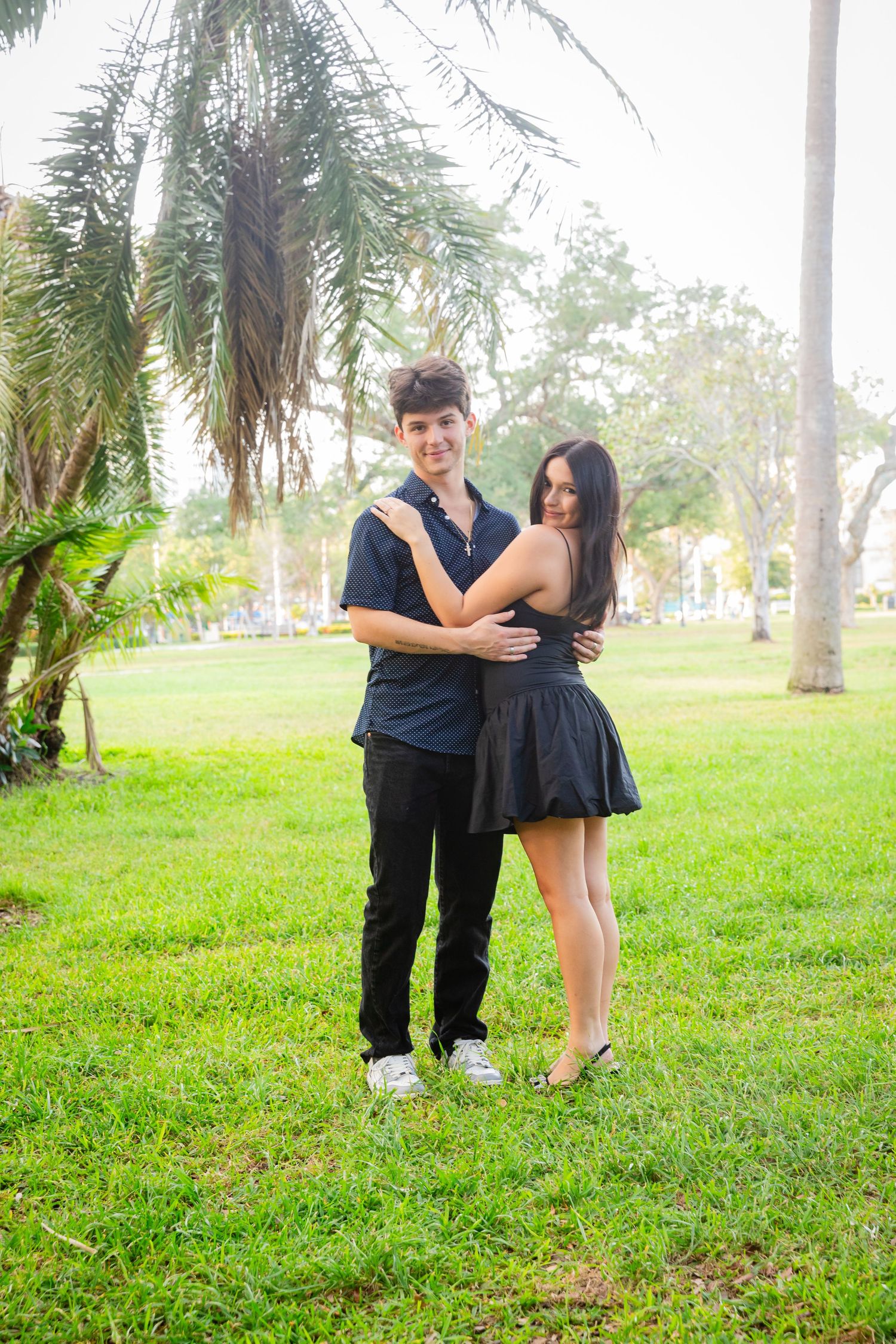 Young couple embracing in a tropical park, woman in black dress and man in dark shirt standing on green grass.
