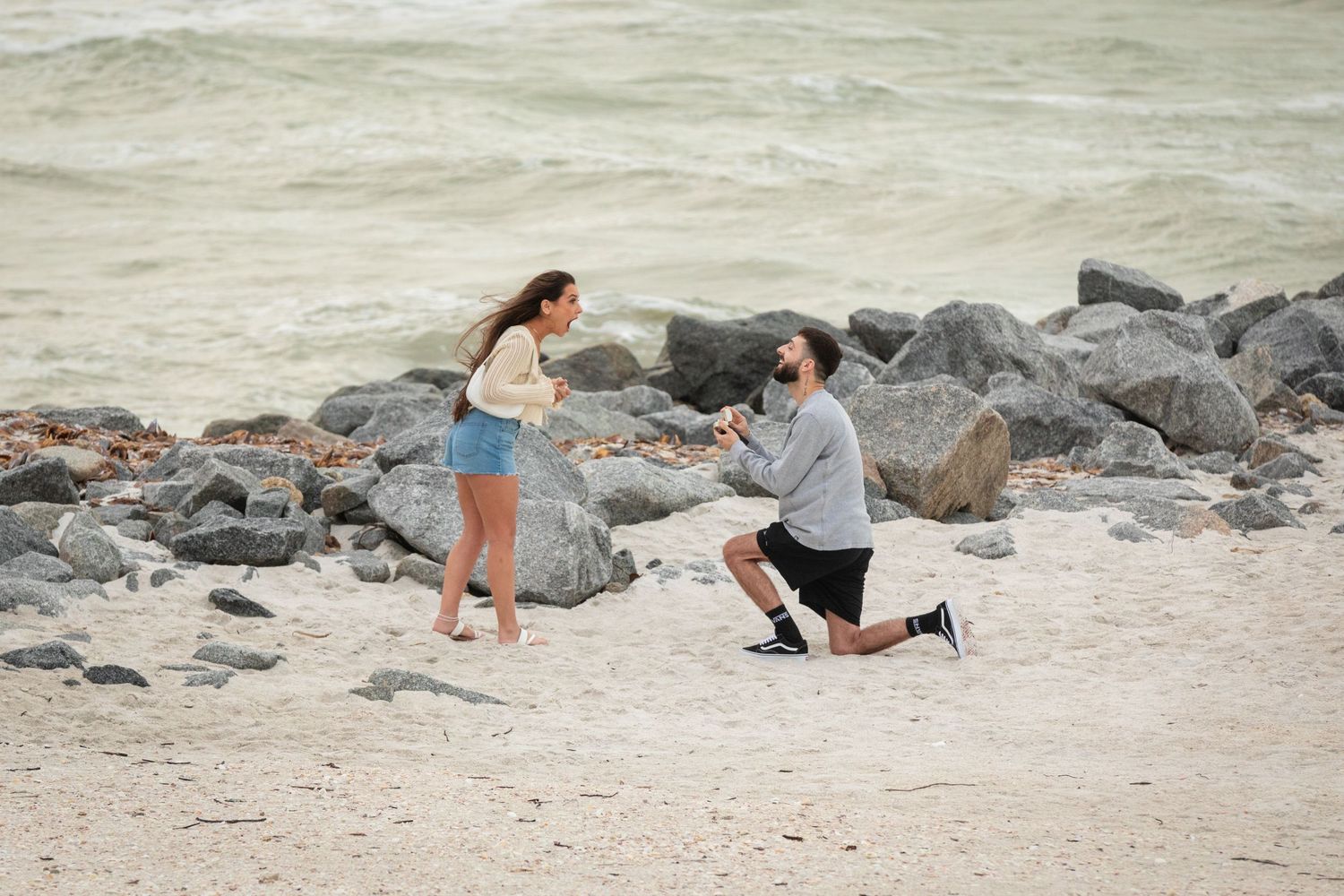 Man proposing to woman on a sandy beach with rocks, down on one knee holding a ring box by the ocean.