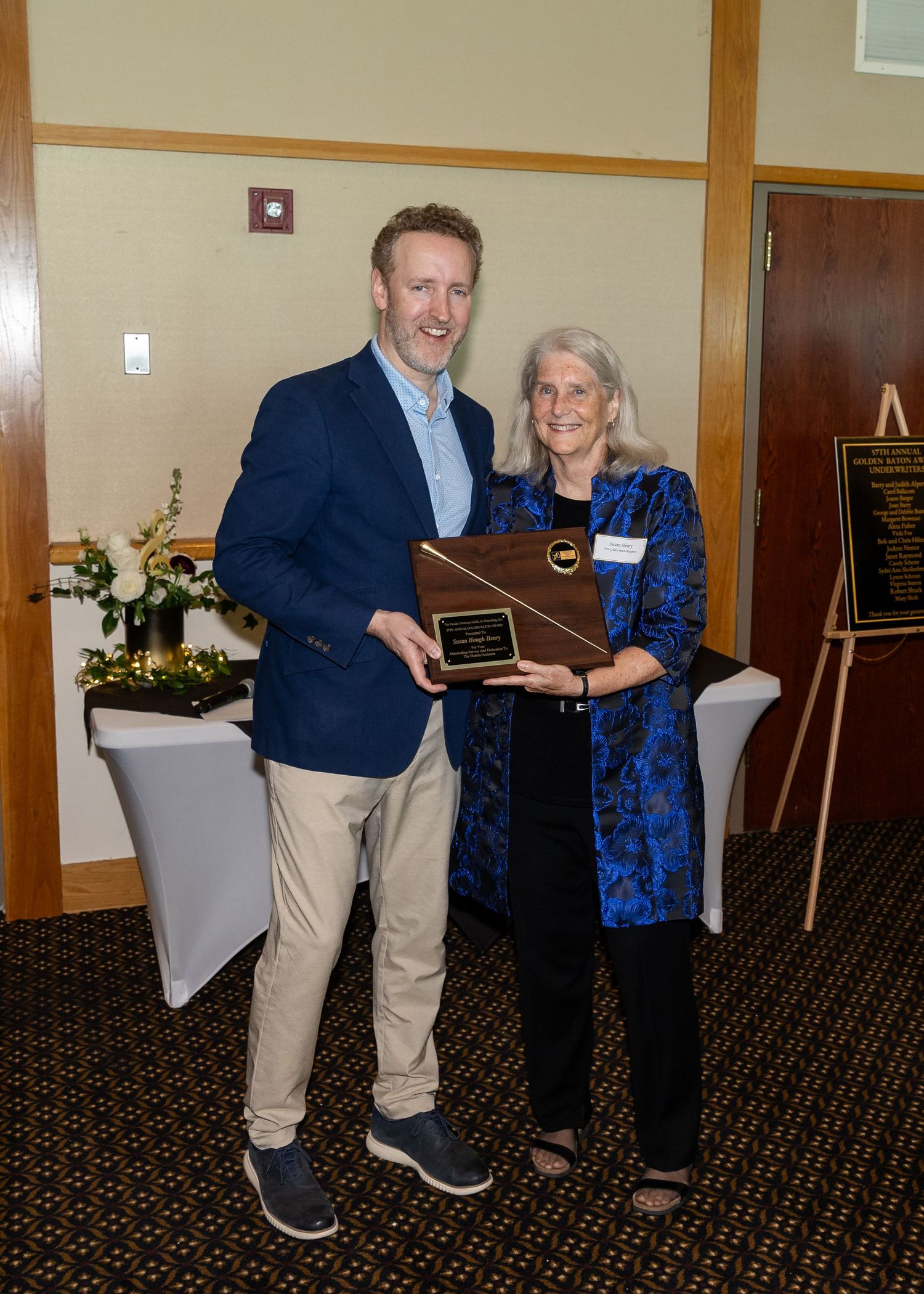 Two people smiling and holding a wooden award plaque at a formal recognition ceremony indoors.
