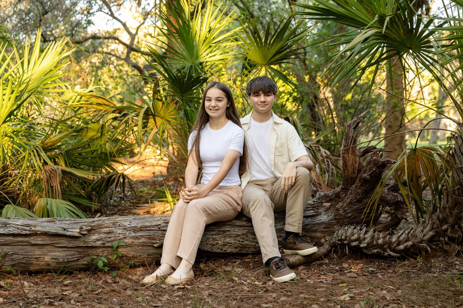 Two teens sit on a fallen log surrounded by lush palm trees in a sunlit forest setting.