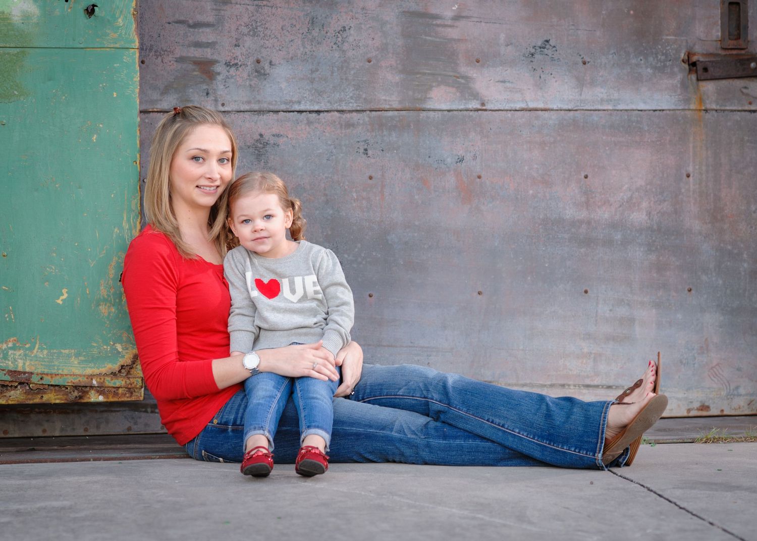 A mother in red top and young daughter in love shirt sit together smiling against a rustic metal wall.