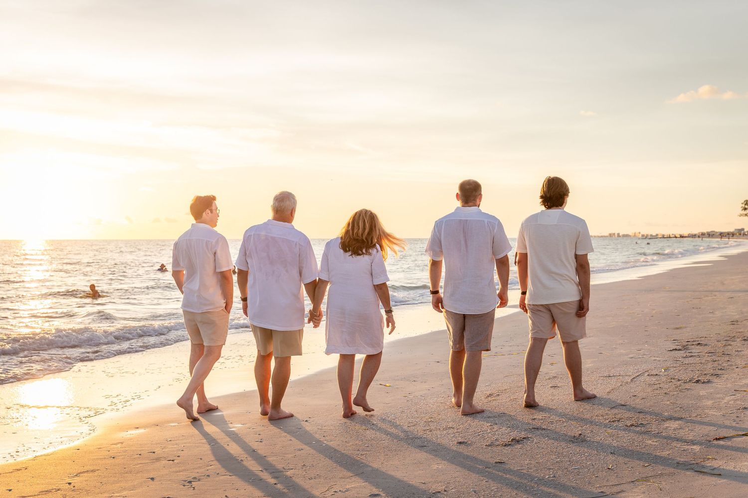 Five people in white shirts walk barefoot on a sandy beach at sunset, viewed from behind.