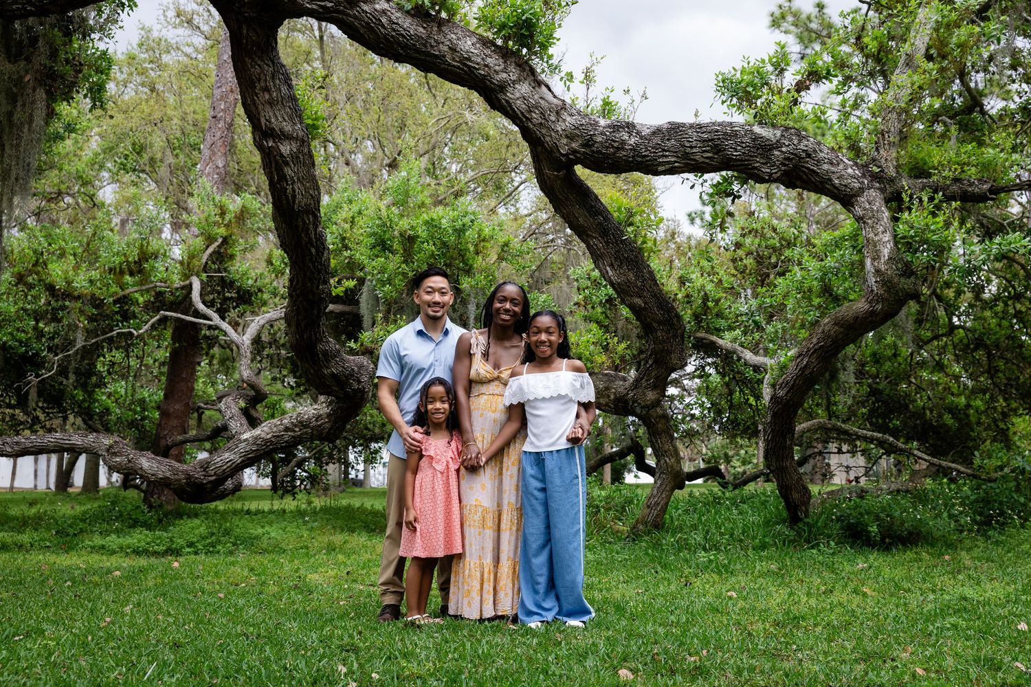 Family of four smiling outdoors under sprawling oak trees on green grass in a park setting.