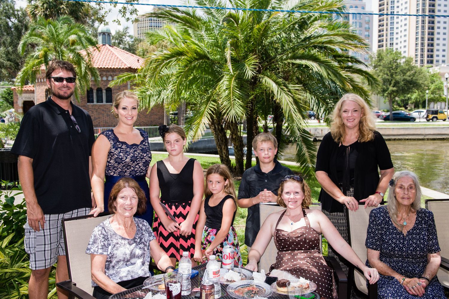 Family group photo outdoors near a pond with palm trees and city buildings in the background on a sunny day.