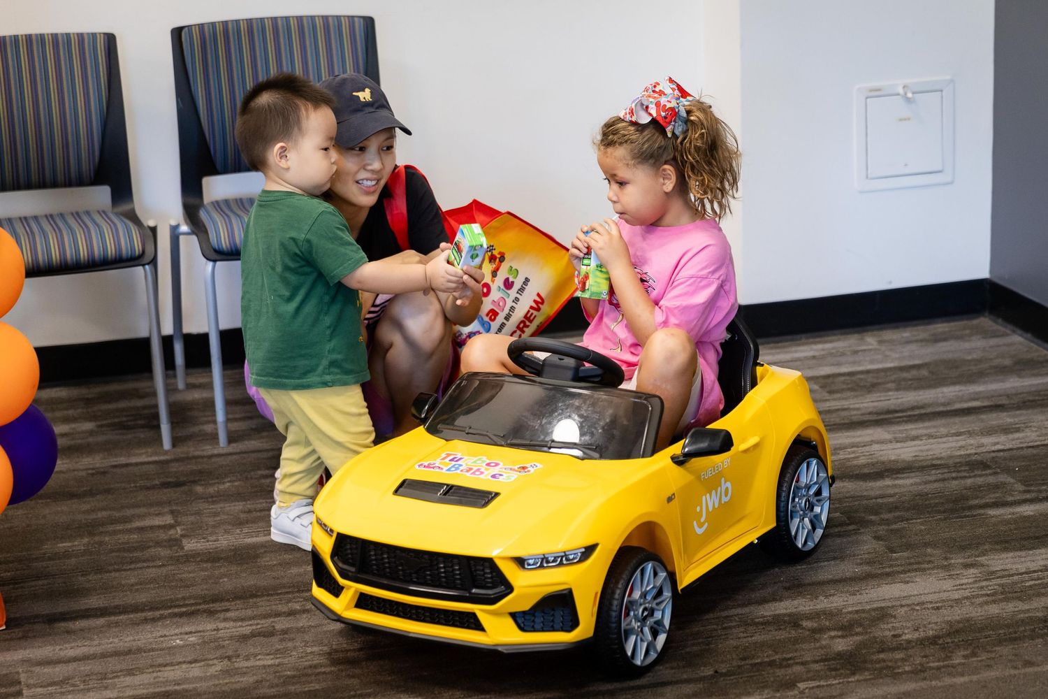 Two children playing with a yellow ride-on toy car indoors, with a woman handing them items near colorful balloons.