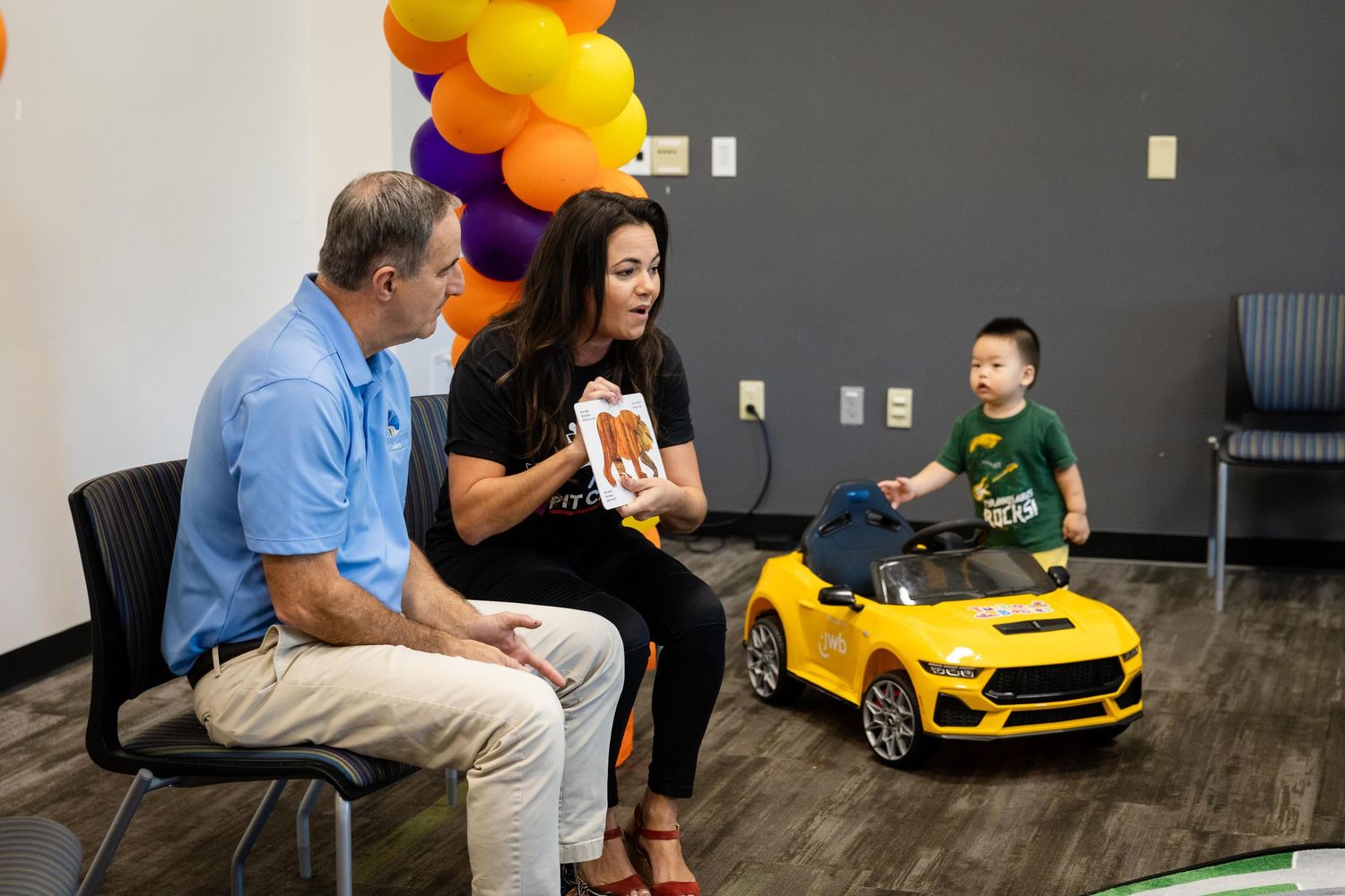 A toddler rides a yellow toy Mustang car while two adults sit nearby at a colorful balloon-decorated indoor celebration.