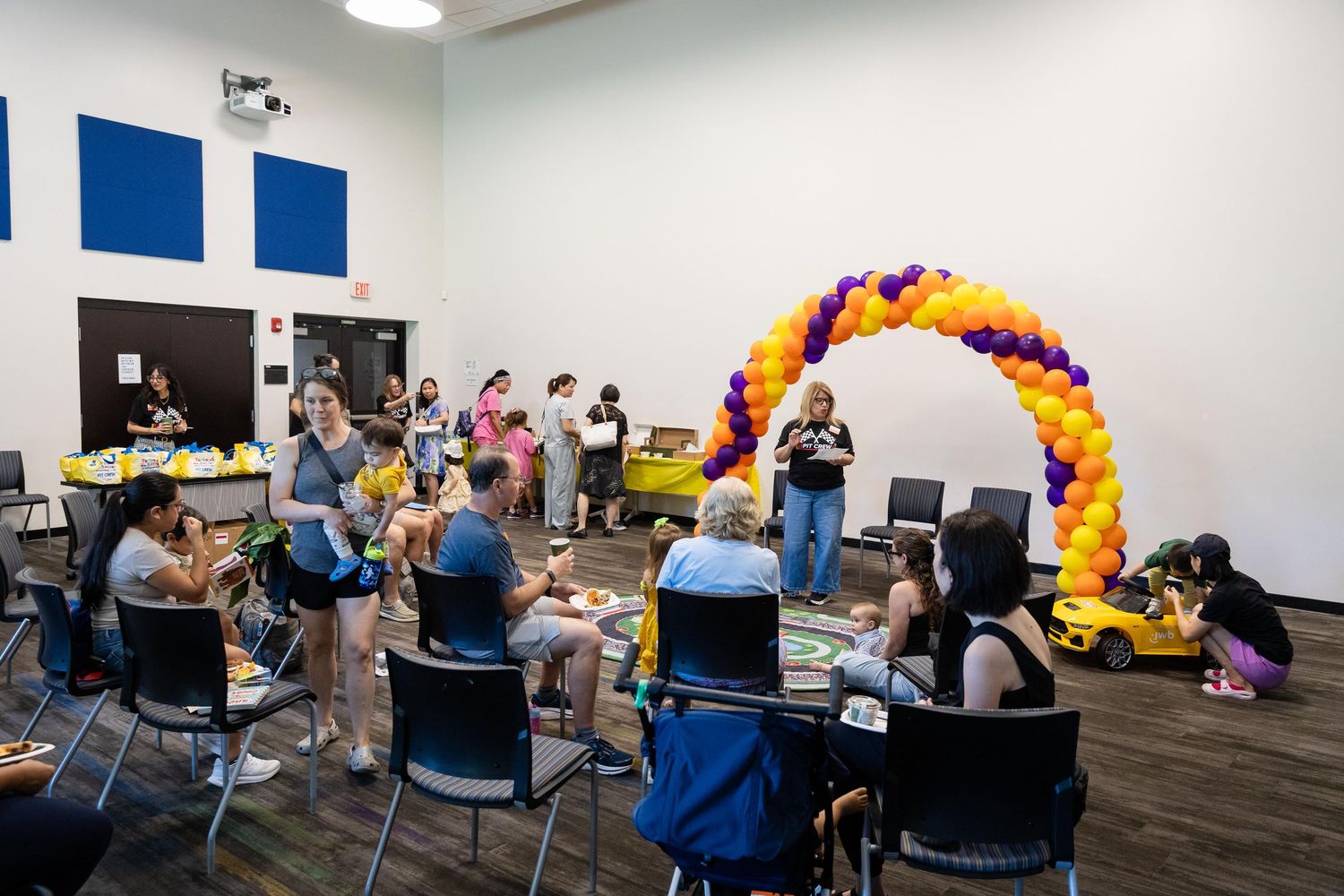 Indoor event with purple and gold balloon arch, attendees seated at tables in a modern community room.