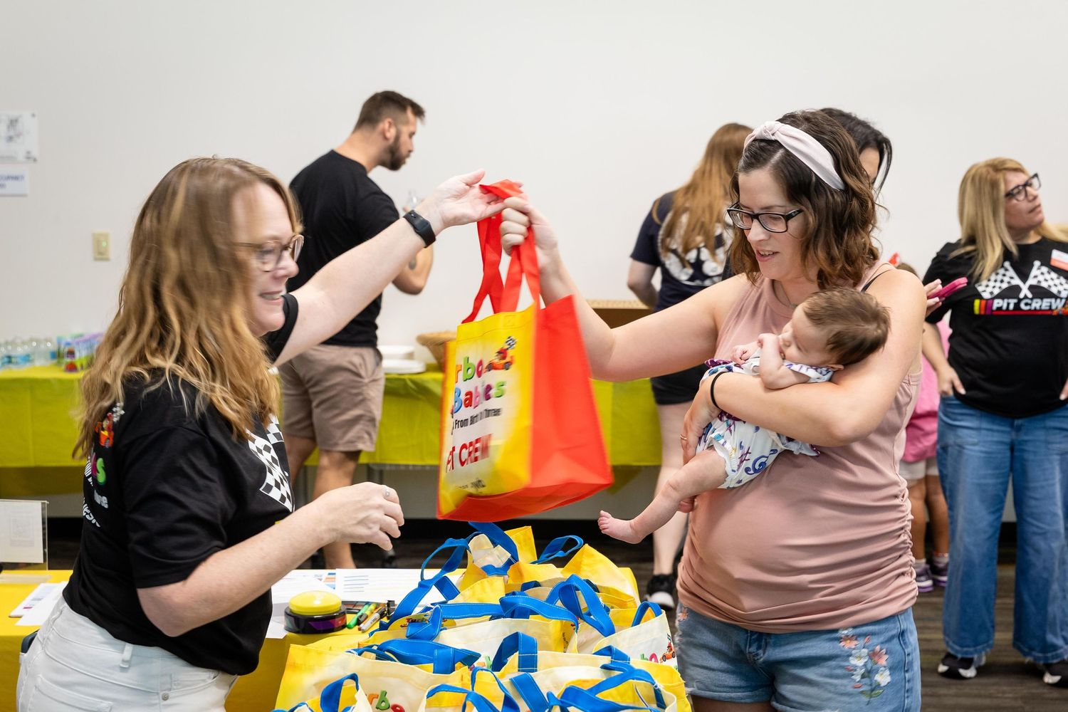 Woman holding baby receives a red gift bag at a community event with people gathered around a table.