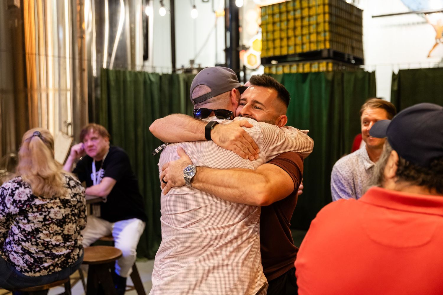 Two men share a warm hug at a casual indoor gathering with friends socializing in the background.