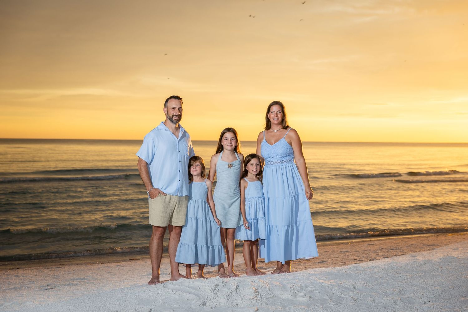 Family of five in matching blue outfits posing on a white sand beach at golden sunset.