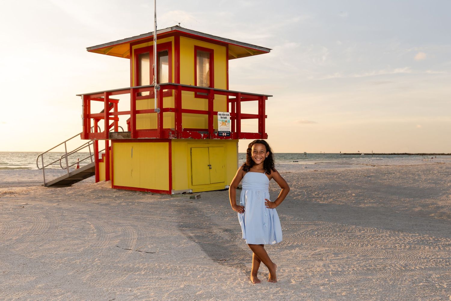 Girl in white dress poses by colorful red and yellow lifeguard stand on sandy beach at sunset.