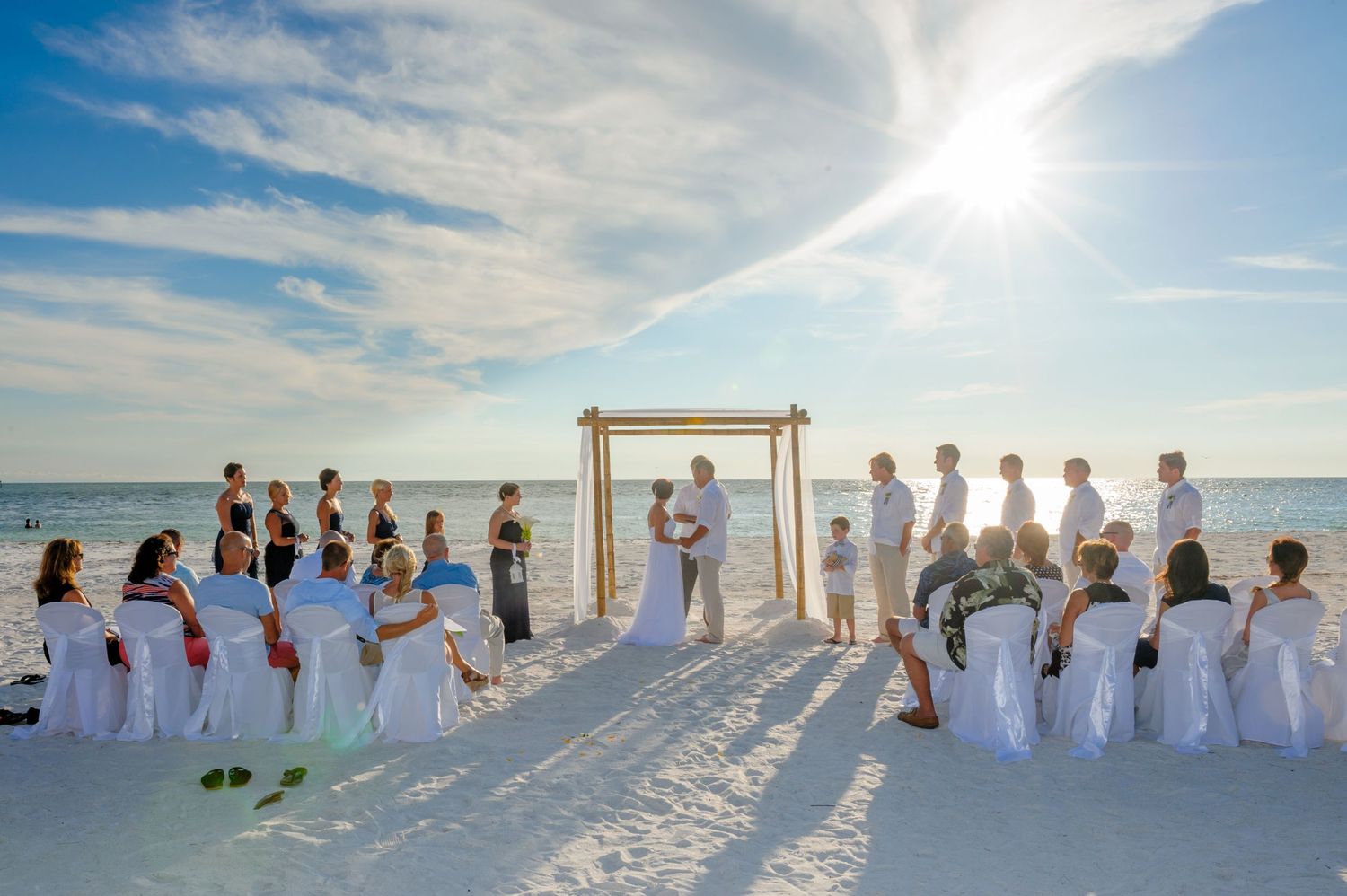 Intimate beach wedding ceremony at sunset with guests seated in white chairs on white sand by ocean.