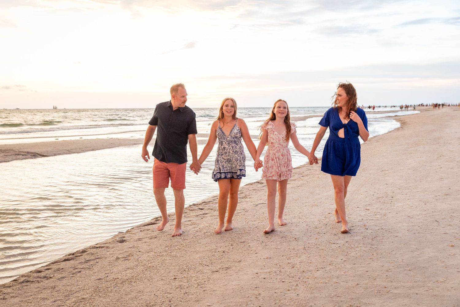 Family of four holding hands walking on a sandy beach at sunset near the ocean shore.