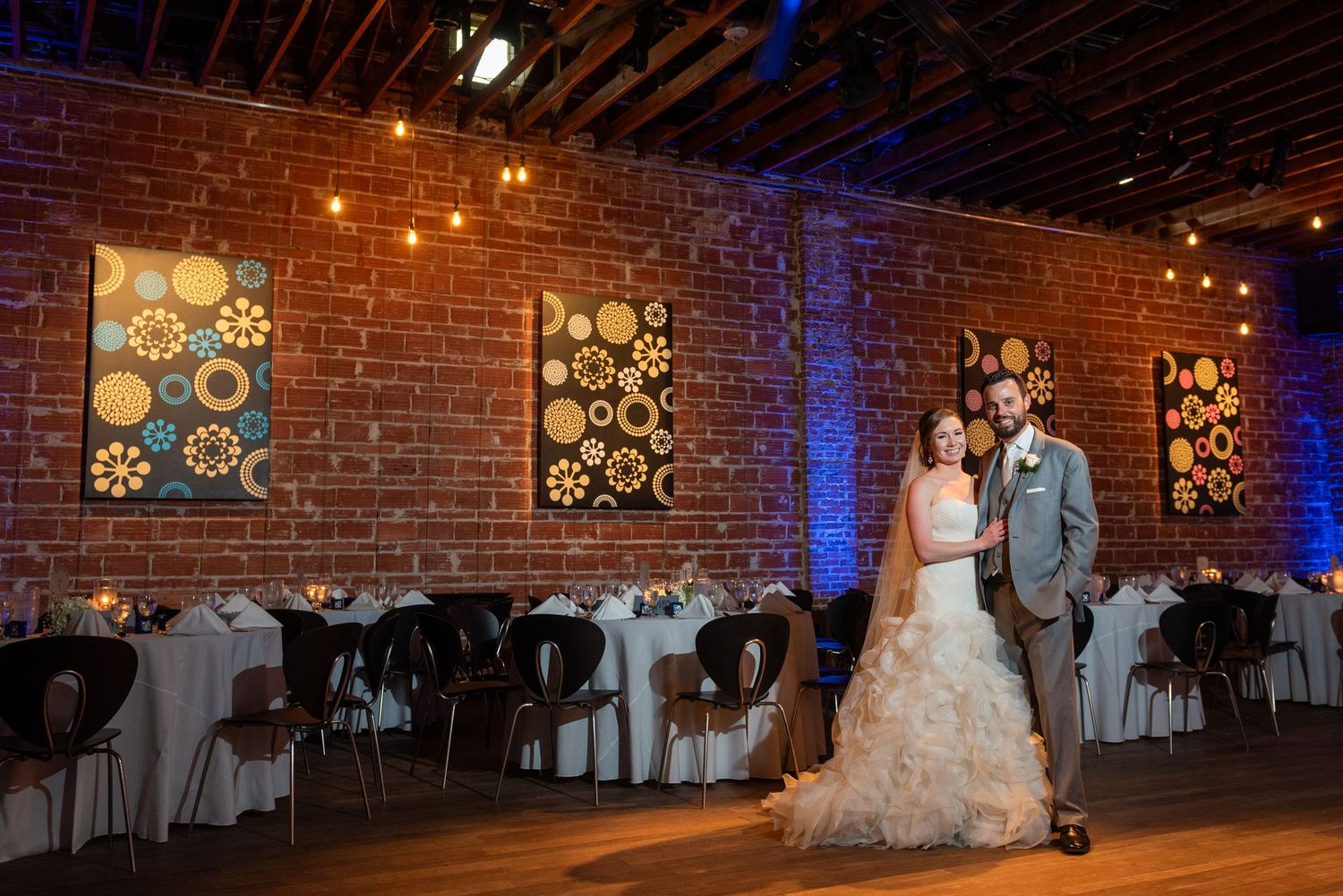Bride and groom pose in elegant rustic wedding venue with exposed brick walls, string lights, and colorful artwork.