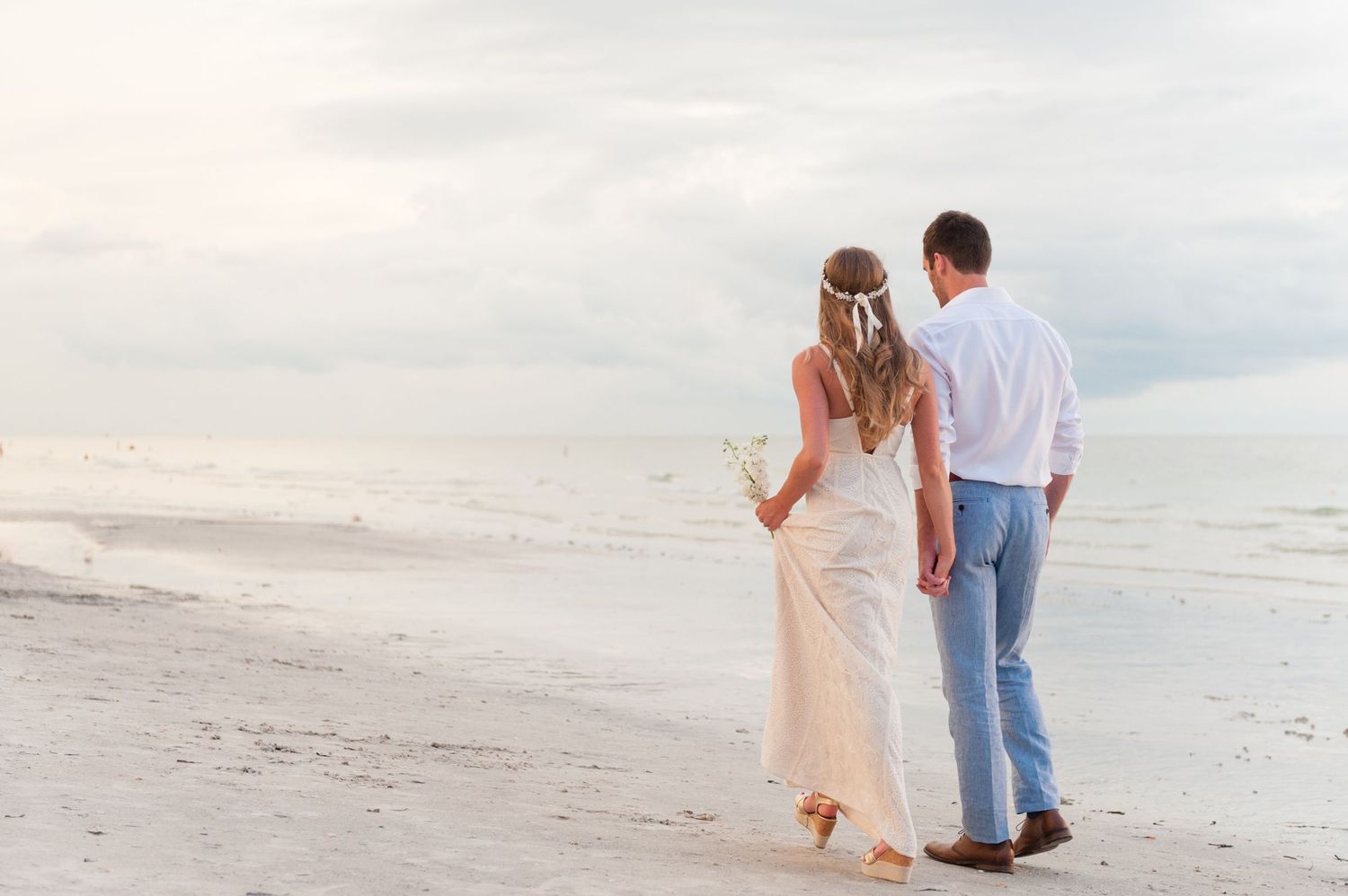 Bride and groom walking hand in hand on a sandy beach at sunset in wedding attire.