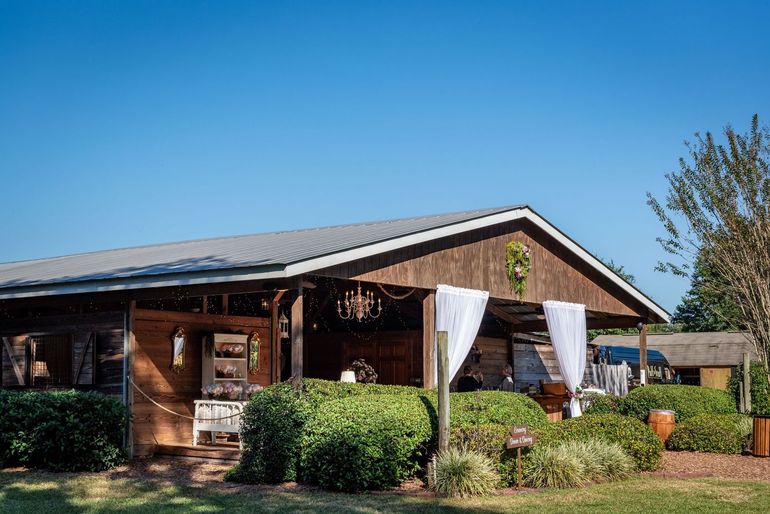 Rustic wooden barn venue with white draping, chandelier, and lush greenery under a clear blue sky.