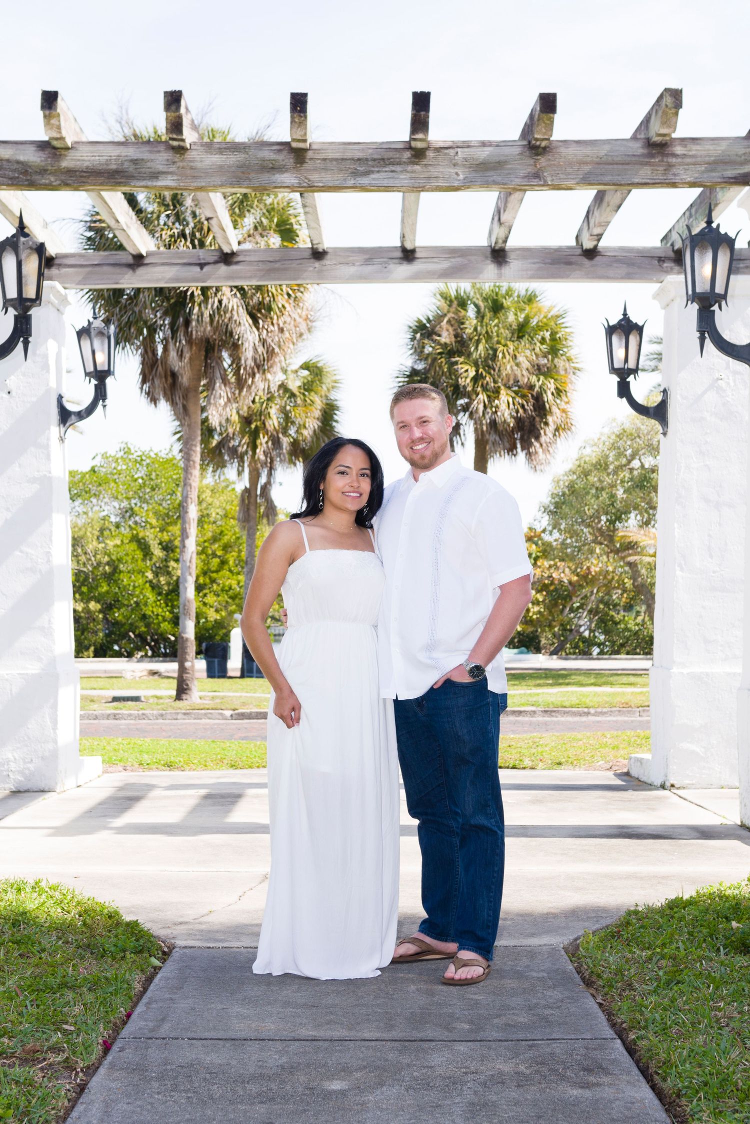 Couple in white outfits posing under a wooden pergola with palm trees in the background on a sunny day.