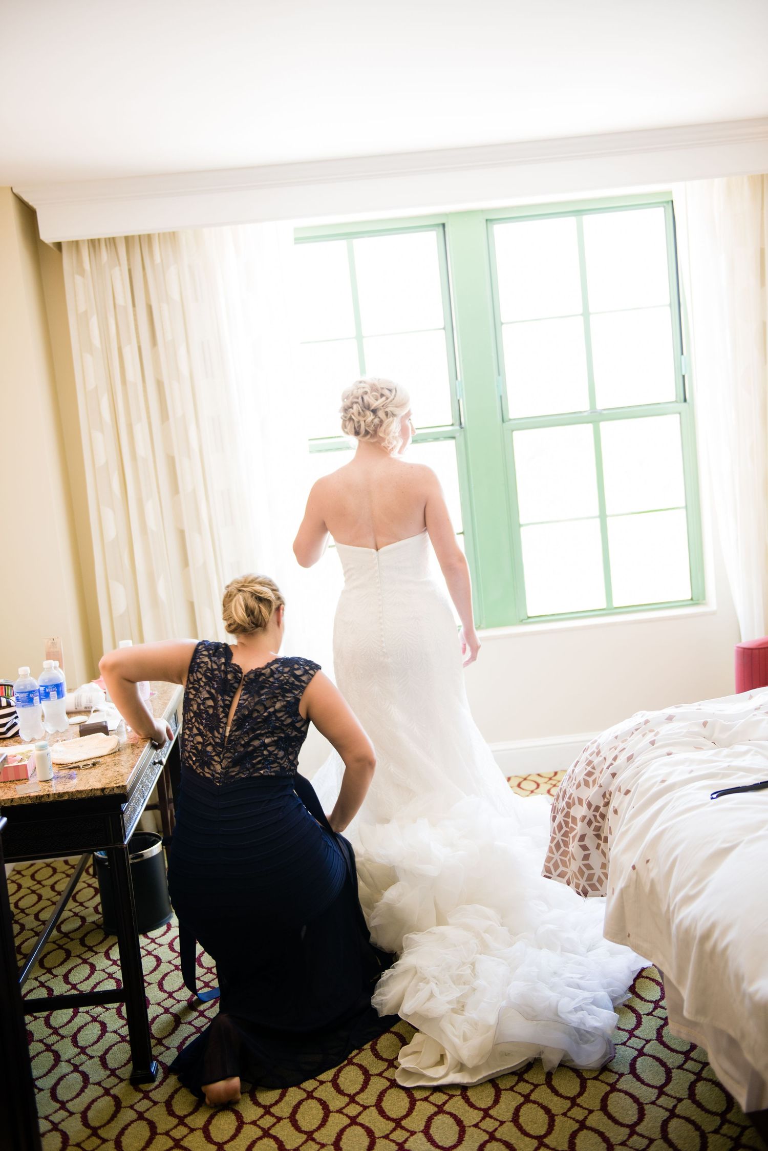 Bride in white wedding gown stands at green-framed window while bridesmaid in navy lace dress sits nearby in hotel room.